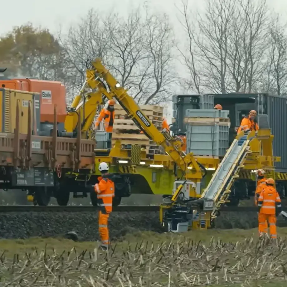 Bauarbeiter in orangefarbener Arbeitskleidung laden Holzpaletten und Baumaterialien auf einen Bauzug.